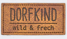 Laden Sie das Bild in den Galerie-Viewer, Detailaufnahme eines geprägten Kunstlederlabels „Dorfkind – wild & frech“ für Hoodies, Taschen und selbstgenähte Accessoires.