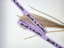 Laden Sie das Bild in den Galerie-Viewer, Baumwoll-Webband „Believe in Unicorns“, 12 mm breit – flieder mit schwarzem schriftzug, längere Bandansicht.
