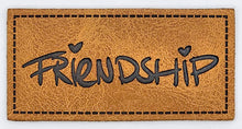 Laden Sie das Bild in den Galerie-Viewer, Detailaufnahme eines geprägten Kunstlederlabels „Friendship“ für Taschen, Hoodies und selbstgenähte Accessoires.
